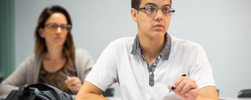 一个学生穿着白色polo衫，戴着眼镜，拿着铅笔向前看，另一个学生在后面，他也在向前看.